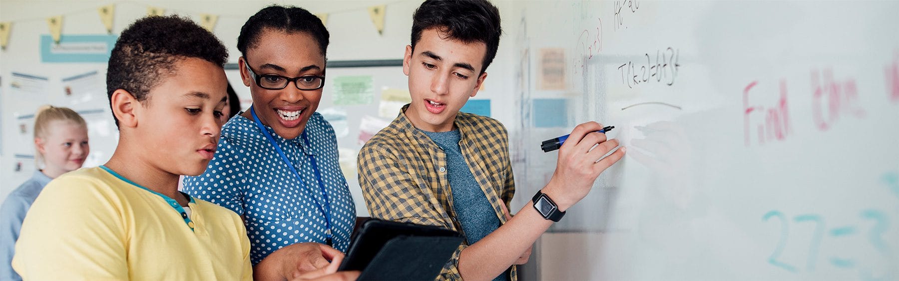 Two young males and a teacher work out a problem on a whiteboard.