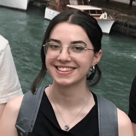 Aurora is a girl with long dark hair that is pulled back. She is wearing round wire glasses, a black tank top and a gray backpack.