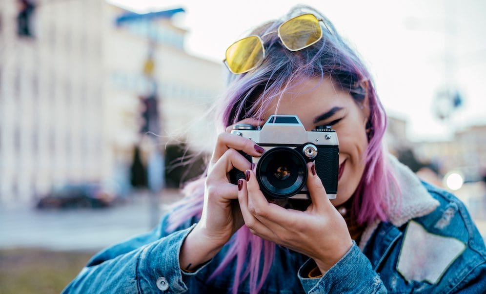 A young woman with pink hair smiles as she points camera toward the viewer