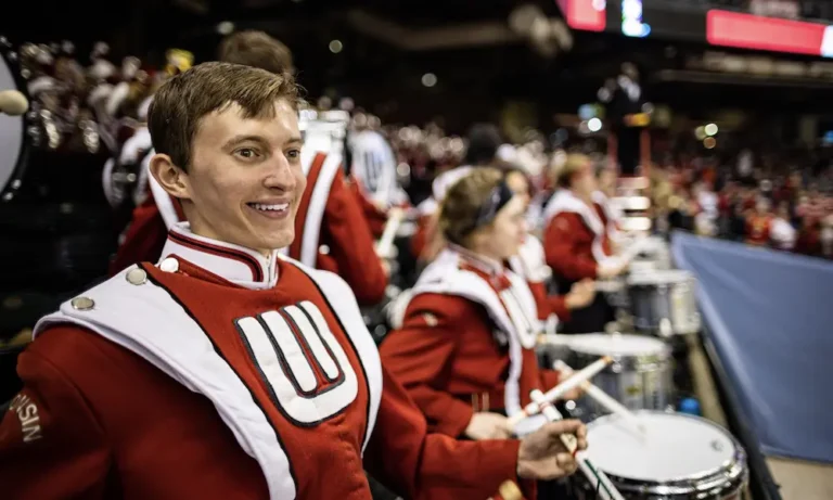 Elliott Frank stands smiling in the foreground. He is wearing a UW Marching Band uniform and has snare drum attached to his front. The the blurred background there are other members of the UW Marching Band, in the stands while watching an unseen event.