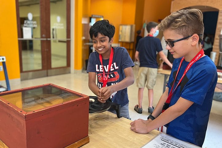 Two boys in blue t-shirts wearing lanyards smile as they watch their experiment