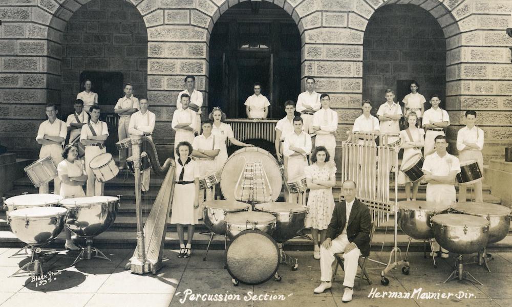 Black-and-white image of 25 young people on the steps of a building. The students are dressed all in white and pose with a selection of percussion instruments. Their instructor sits in a chair at the bottom of the steps in front of the students.