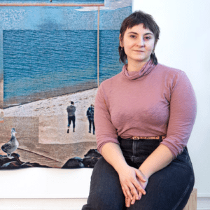 Annmarie Suglio sits in front of work of art depicting two figures on a pier with ocean in background