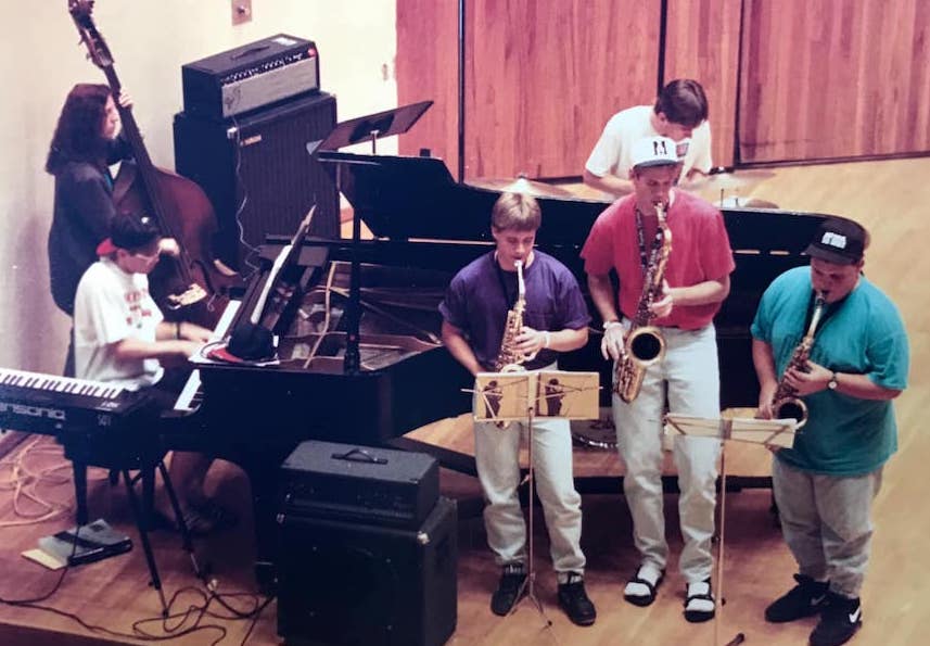Six young musicians play together. A young woman plays stand-up bass. One young man plays piano another plays drum kit. Three young men stand in the foreground playing saxophones.