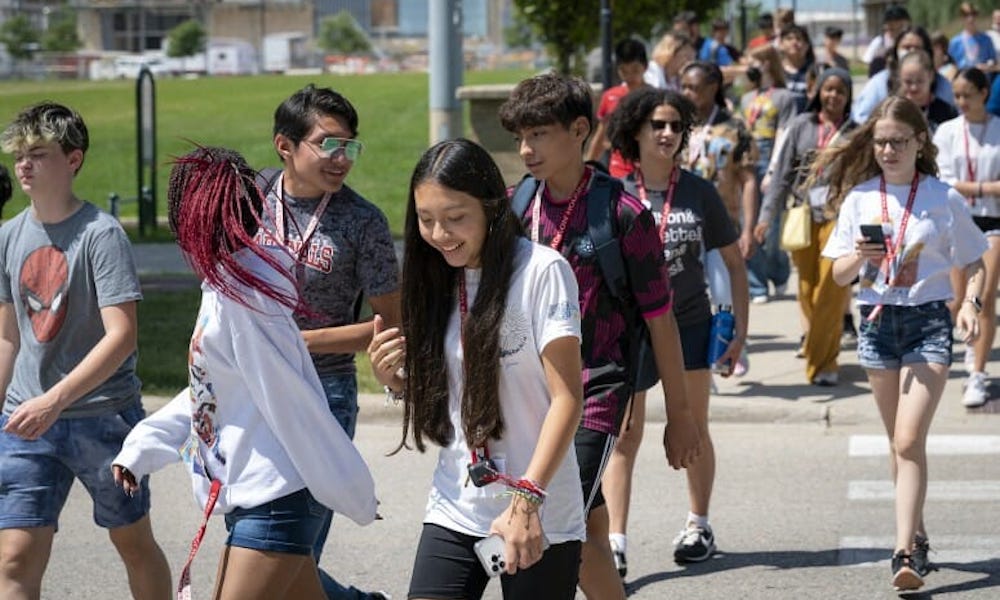 A diverse group of middle school students walks down a sidewalk on campus smiling and talking. More students appear in the background.