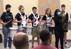 Six high school students stand performing percussion instruments in front of an audience. Five students play the snare drum and one plays the cymbals.