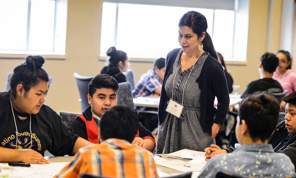 A female instructors walks between tables of Latinx middle school students, encouraging their work during UW–Madison's Latino Youth Summit.