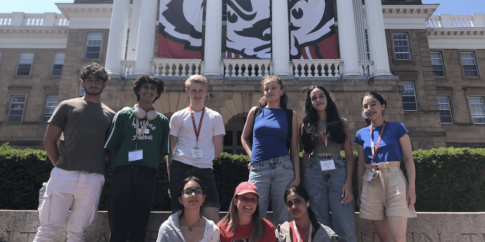 Nine international high school students pose in front of UW–Madison's Bascom Hall. A large banner of Bucky Badger is hanging behind the building's columns