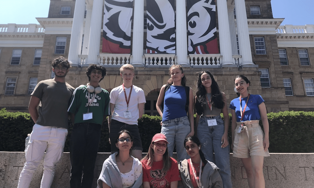 Nine international high school students pose in front of UW–Madison's Bascom Hall. A large banner of Bucky Badger is hanging behind the building's columns