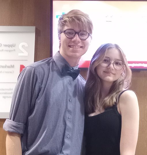 Jacob and Willow pose together. Jacob is tall with blonde hair and is wearing a blue shirt, bow tie and glasses. Willow has long blonde hair and wears a sleeveless black dress and glasses. 