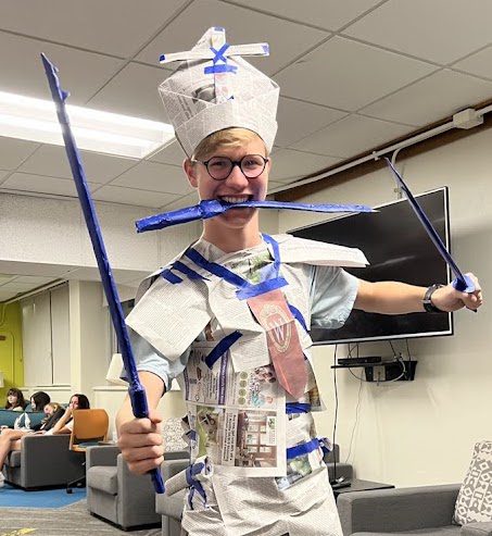 Jacob as an RA, dressed in handmade newspaper hat and clothing with holding two paper "swords" and a "knife" in his teeth, smiles at the camera. He has blonde hair and wears glasses.
