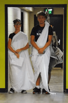 Two teen boys stand in a doorway dressed in white toga-style sheets with dark blue t-shoes and tennis shoes showing beneath. A person dressed in newspaper is barely visible behind them.