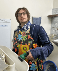 Ian Vailliencourt looks at the camera as he stands before a large sink. He wears a colorful apron, blue shirt and glasses and his holding a water sprayer.