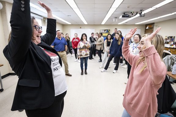 Lisa Barker stands smiling with hands raised facing a student who is mimicking her posture and expression. In the background, others stand observing this activity, smiling.