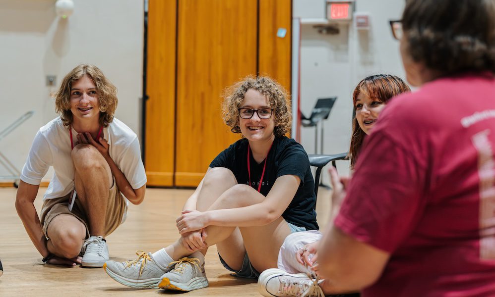 Three high school students sit on the floor, smiling as they face their instructor, whose back is in the foreground. Students are wearing t-shirts and shorts.